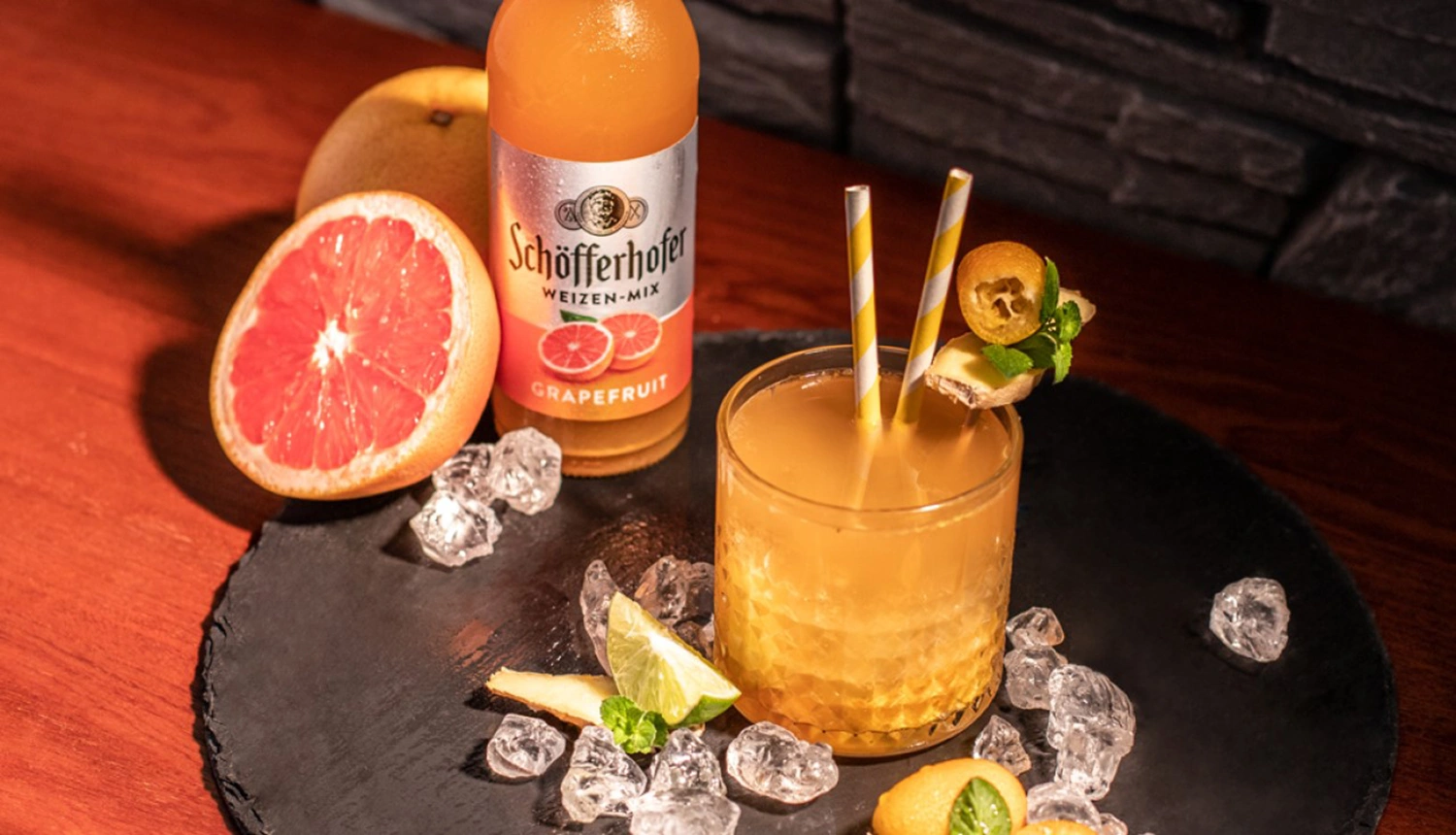 Ein Getränk steht auf einem Tisch neben geschnittener Grapefruit und Eiswürfeln. Zwei gestreifte Strohhalme ragen aus dem Glas.