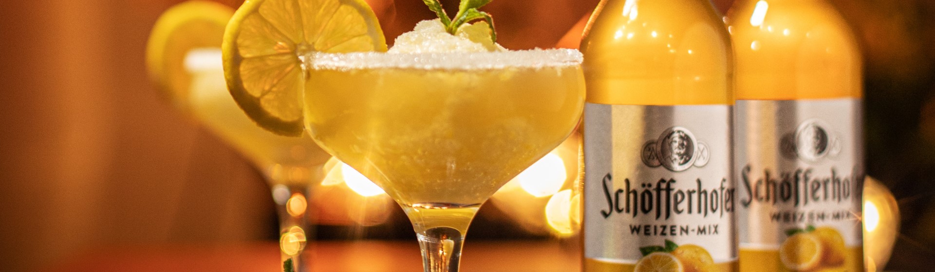 Ein Cocktailglas mit Zuckerrand und Limettenscheibe steht im Vordergrund, daneben sind Getränkeflaschen.
