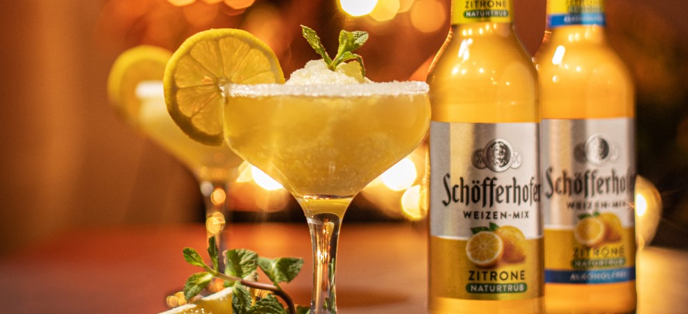 Ein Cocktailglas mit Zuckerrand und Limettenscheibe steht im Vordergrund, daneben sind Getränkeflaschen.