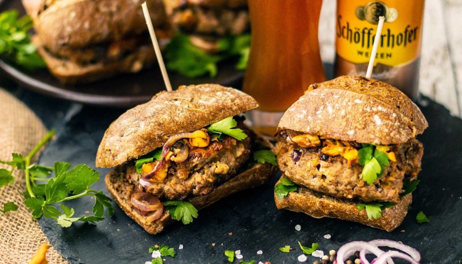 Zwei Vollkornbrötchen mit Bier-Frikadellen, Petersilie, Pilzen und Zwiebeln. Im Hintergrund ein Glas Hefeweizen.
