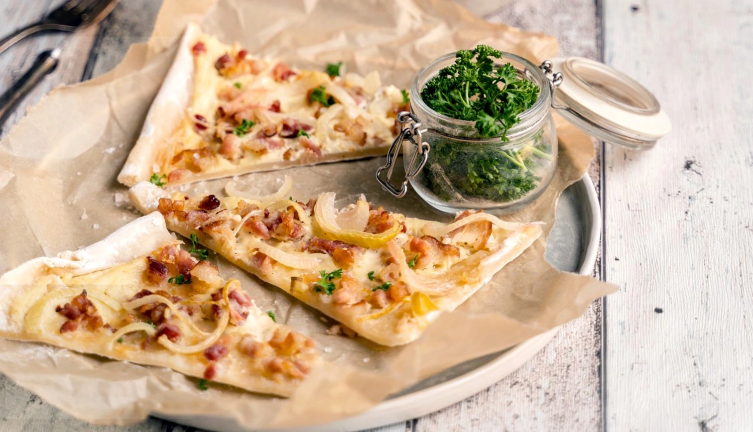 Ein Teller mit drei Stücken dünner Flammkuchen belegt mit Zwiebeln und Speck, daneben ein Glas mit Kräutern.
