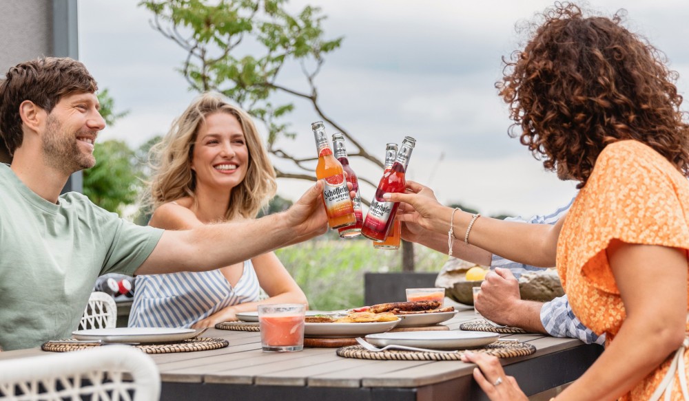 Drei Personen stoßen mit Getränken an einem Gartentisch an. Sie lächeln und genießen das Essen.